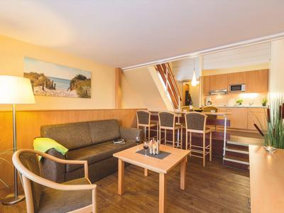 Helles Wohnzimmer mit braunem Sofa, Holzstuhl, Stehlampe und Bartresen mit drei Hockern.
