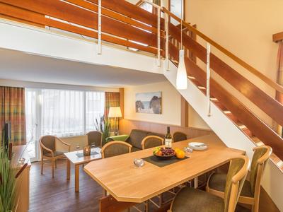 Helles Wohnzimmer mit Holzmöbeln, Esstisch, Sofa, Sessel und offener Holztreppe zur Empore.