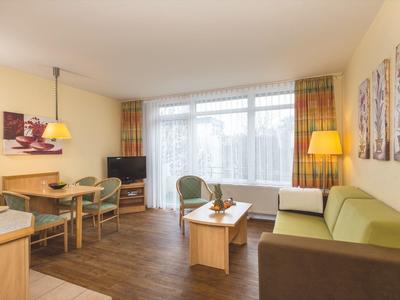 Helles Wohnzimmer mit grünem Sofa, Holztisch, Essbereich, Fernseher und großen Fenstern mit Vorhängen.