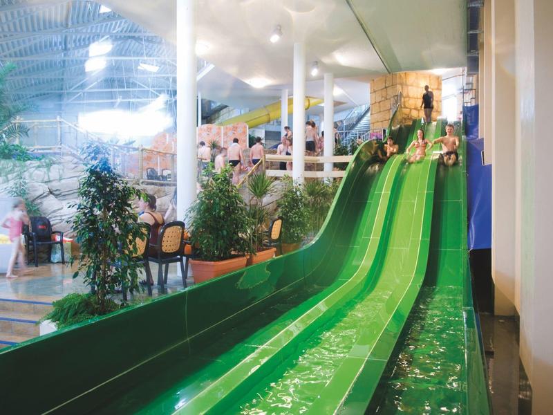 Grüne Wasserrutschen in einer hellen Indoor-Wasseranlage mit Pflanzen und Sitzbereich.