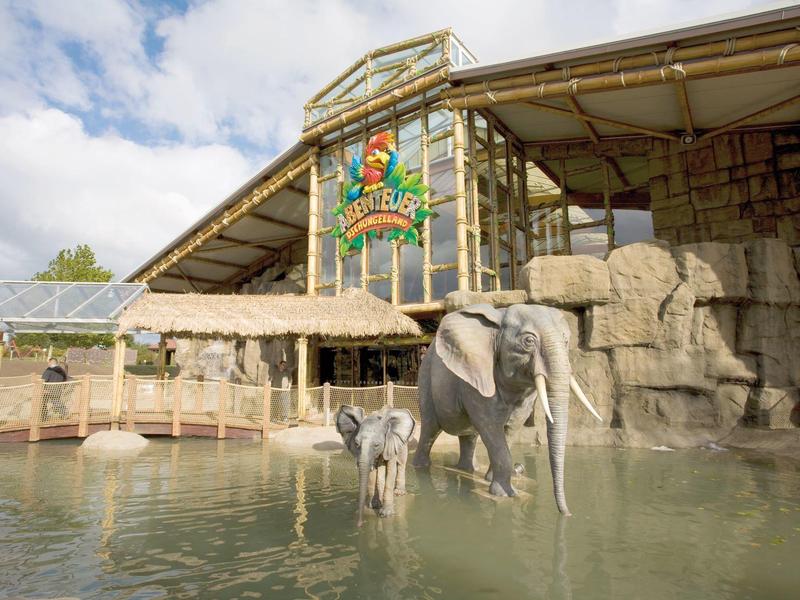 Elefant mit Jungtier steht im Wasserbecken vor einer großen, offenen Halle mit Felsen und Logo.