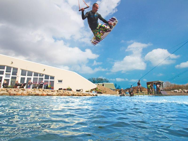 Ein Kitesurfer springt hoch über blauem Wasser vor blauem Himmel und Strandkulisse.