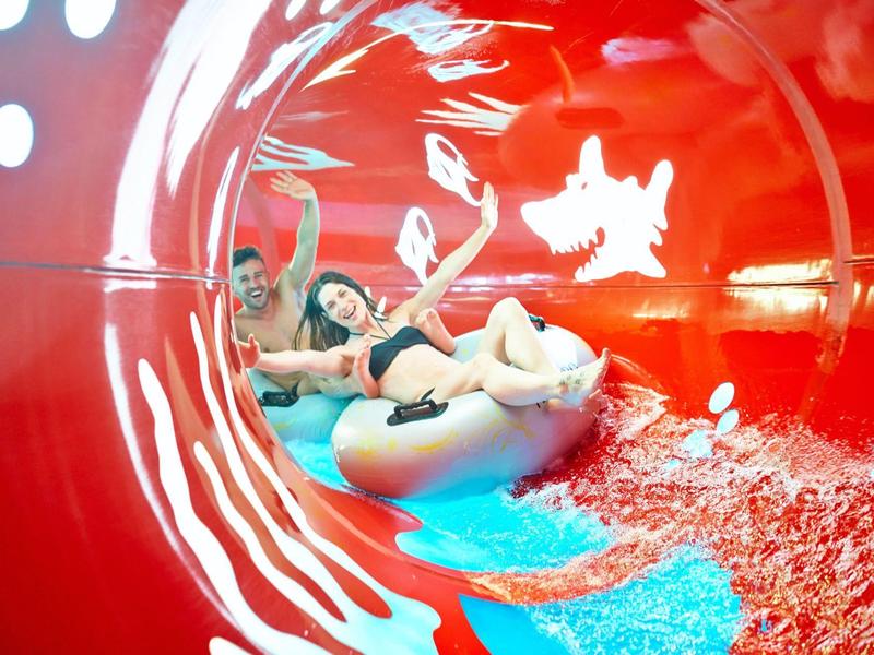 Zwei Personen rutschen lachend in einem roten Wasserrutschen-Tunnel auf einer blauen Schwimmhilfe.