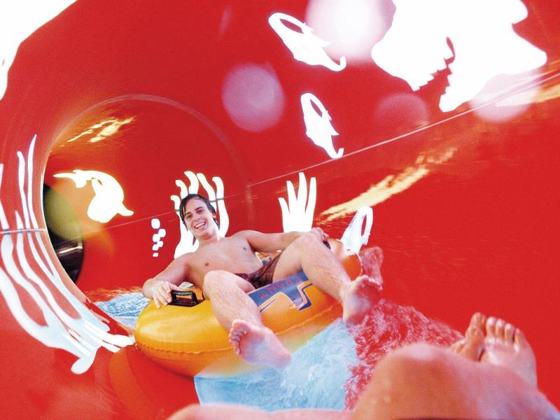 Zwei Personen rutschen in einem roten Wasserrutschen-Tunnel mit Spritzwasser und heller Beleuchtung.