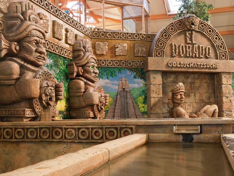 Farbenfrohe Detailwand mit Mayastatuen und Tempelreliefs über einem Wasserkanal im Indoor-Bereich.