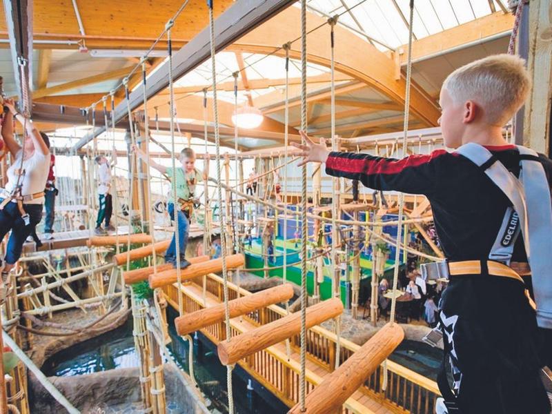 Kind klettert in einer Indoor-Hochseilgarten-Anlage mit Holzplattformen und Sicherheitsgurt.