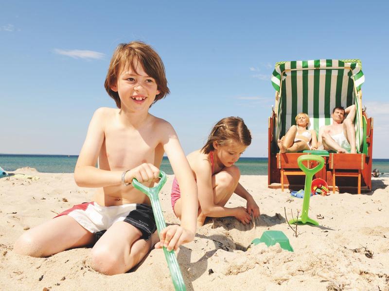 Zwei Kinder spielen im Sand am Strand, während zwei Erwachsene im Strandkorb sitzen.