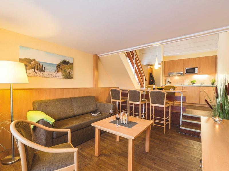 Helles Wohnzimmer mit braunem Sofa, Holzstuhl, Stehlampe und Bartresen mit drei Hockern.