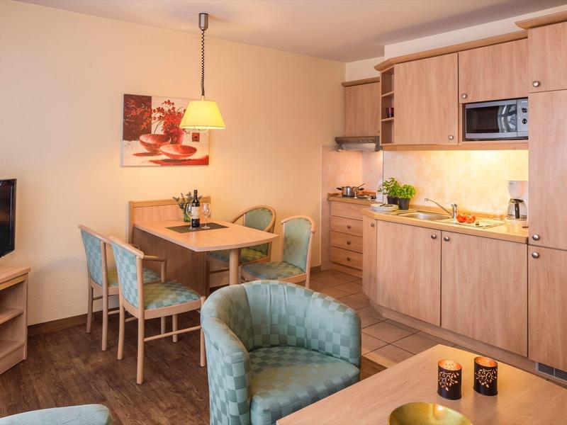 Helle kleine Küche mit Holzschränken, Esstisch, Stühlen und gemütlichem Sessel in warmem Licht.