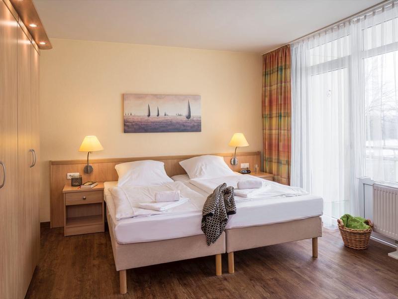 Helles Schlafzimmer mit Doppelbett, Holzboden, Nachttischen, großer Gardine und Wandbild.