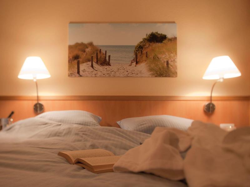 Beleuchtetes Doppelbett mit weißen Bettdecken, offenem Buch und Dünenstrandbild an Wand.