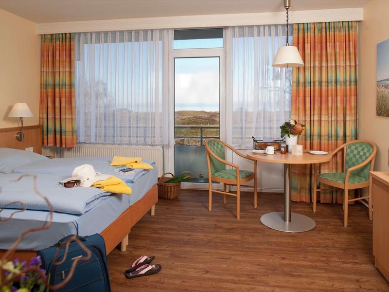 Gemütliches Hotelzimmer mit Doppelbett, Balkon, Tisch mit Stühlen und hellen Vorhängen.
