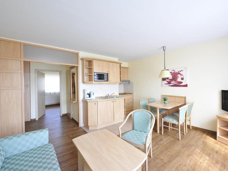 Helles Wohnzimmer mit Holzfußboden, Sitzecke, Esstisch, Küchenzeile und Fernseher neben Fenster.