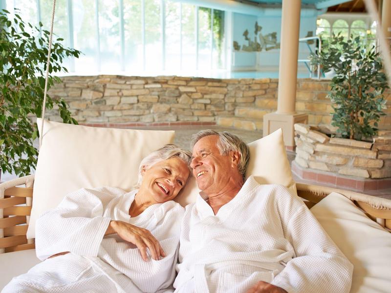 Älteres Paar entspannt sich lachend auf einer hellen, bequemen Couch in gemütlichem, hellem Raum.