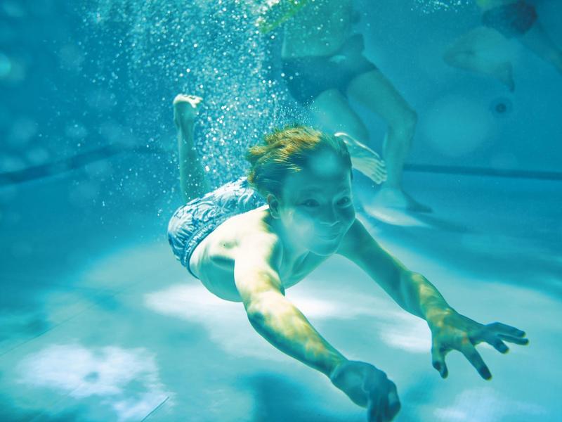 Mann taucht im klaren Wasser, ausgestreckter Arm, Lichtreflexe und Luftblasen um ihn herum.