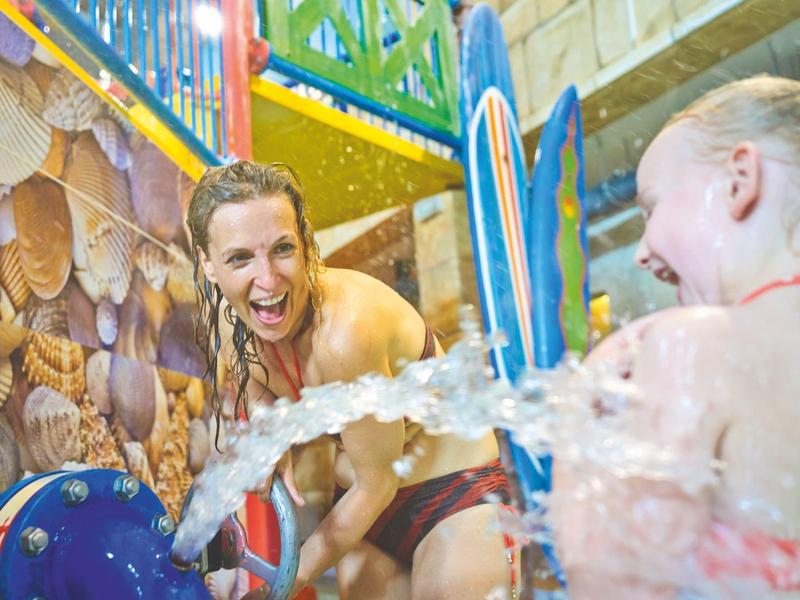Frau und Mädchen in Badebekleidung spielen lachend mit Wasserstrahl im bunten Indoor-Wasserspielbereich.