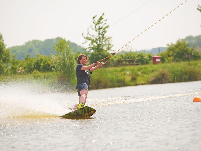 Mann wakeboardet auf ruhigem See, umgeben von grünen Bäumen und bewölktem Himmel.