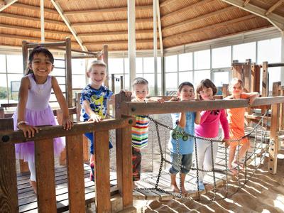 Sechs Kinder spielen in einem sonnendurchfluteten, holzgebauten Indoor-Spielplatz.