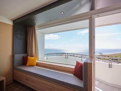 Sitzbereich am Fenster mit Aussicht auf das Meer und Balkon im Hotelzimmer.
