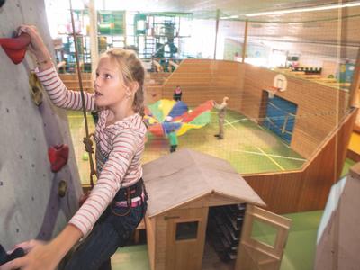 Ein Mädchen klettert an einer Kletterwand in einer Indoor-Freizeitanlage mit Spielbereich.