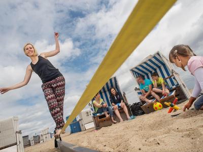Frau balanciert auf Slackline am Strand, Zuschauer und Kinder sitzen im Sand.