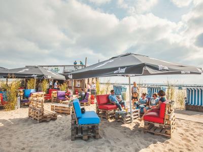 Strandbar mit bunten Sitzmöbeln unter großen Sonnenschirmen und Sandboden.