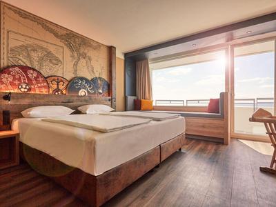 Hotelzimmer mit Doppelbett, großem Wandbild und Fensterbank mit Sitzmöglichkeit und Meeresblick.