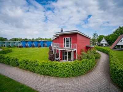 Rotes Ferienhaus mit Terrasse, umgeben von grünen Hecken und gepflasterten Wegen, bei bewölktem Himmel.