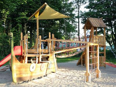 Ein hölzerner Spielplatz mit Rutschen und Kletterbrücken auf Sandboden, umgeben von Bäumen.