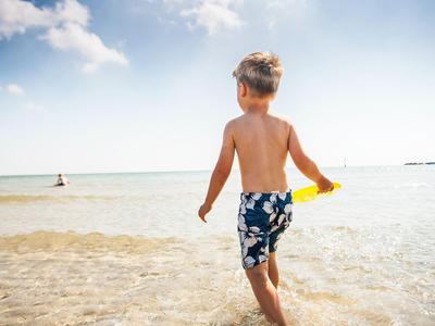 Ein Junge in Badehose geht am flachen Strand ins Wasser und hält eine gelbe Schaufel.