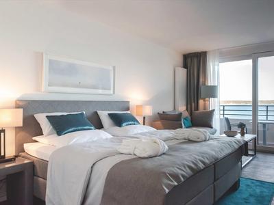 Modernes Schlafzimmer mit grauem Bett, blauen Kissen, zwei Nachttischlampen und Balkon mit Meerblick.