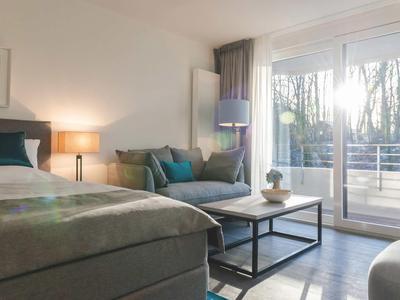 Helles Schlafzimmer mit großem Bett, grauer Couch, Holzboden und bodentiefen Fenstern mit Balkon.