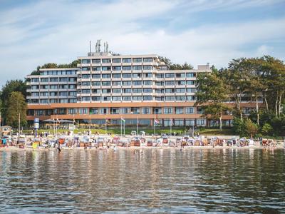 Großes Hotelgebäude mit Balkonreihen, Strandkörben und Wasser im Vordergrund bei klarem Himmel.