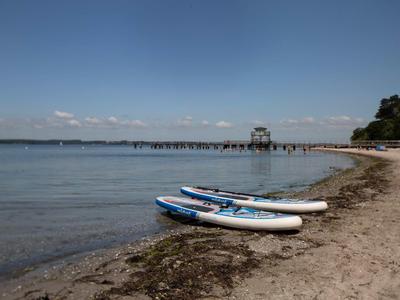 Strand mit zwei Stand-Up-Paddleboards am Ufer und Pier im Hintergrund bei klarem Himmel.