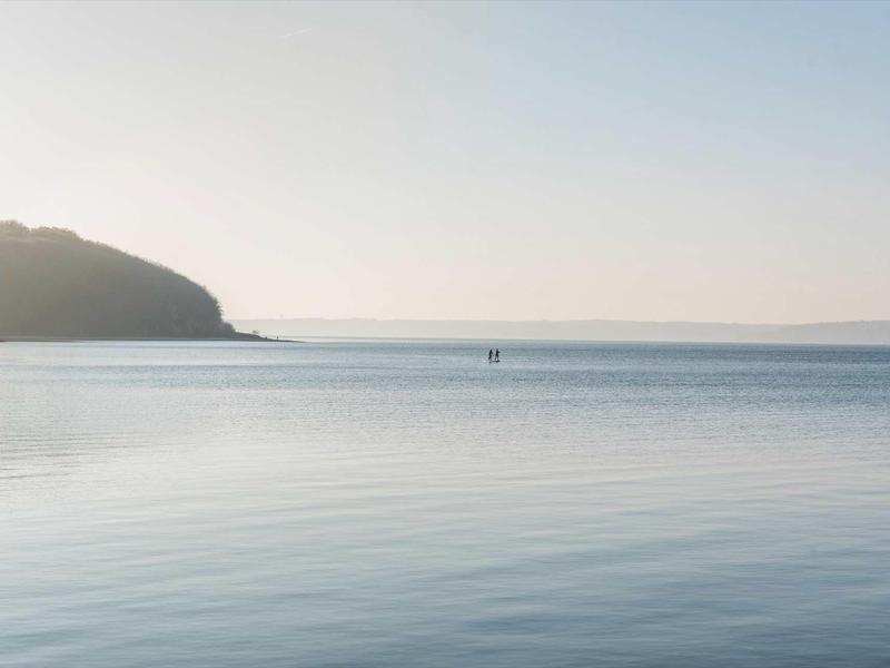 Weites, ruhiges Meer mit sanften Wellen, weiter Horizont und bewaldeter Hügel links im Bild.