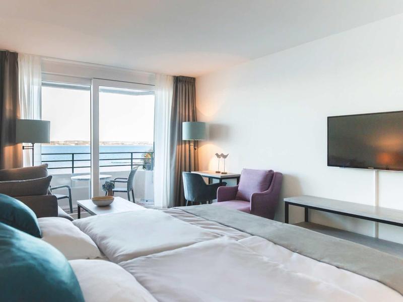 Modernes Hotelzimmer mit Doppelbett, Sitzbereich, Flachbildfernseher und großem Fenster mit Balkon.