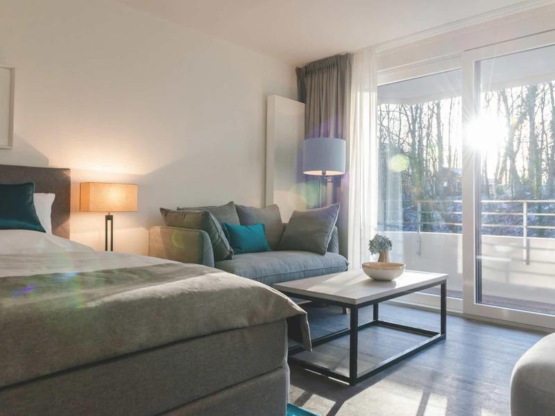 Helles Schlafzimmer mit großem Bett, grauer Couch, Holzboden und bodentiefen Fenstern mit Balkon.