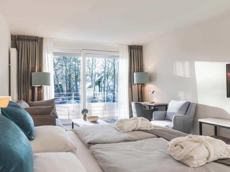 Helles, modernes Schlafzimmer mit großem Bett, blauen Kissen, Sesseln und Balkon mit Glasfront.