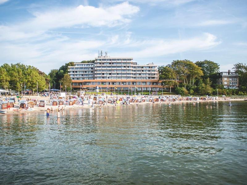 Strand mit Menschen, Bäumen und einem großen Hotelgebäude am Ufer unter blauem Himmel.
