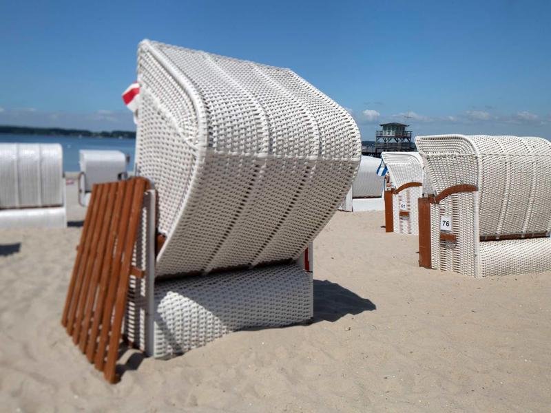 Weiße Strandkörbe aus Geflecht stehen auf hellem Sandstrand unter blauem Himmel.