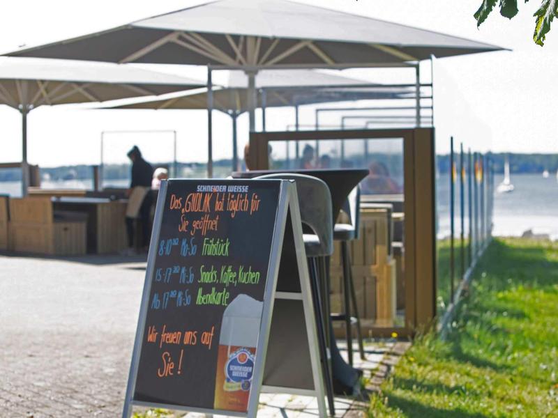 Straßenaufsteller vor Café mit Sonnenschirmen, Blick auf Wasser und grünen Rasen am sonnigen Tag.