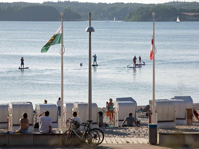 Menschen sitzen an einer Promenade mit Strandkörben, Segelboote und Paddler sind auf dem Wasser zu sehen.