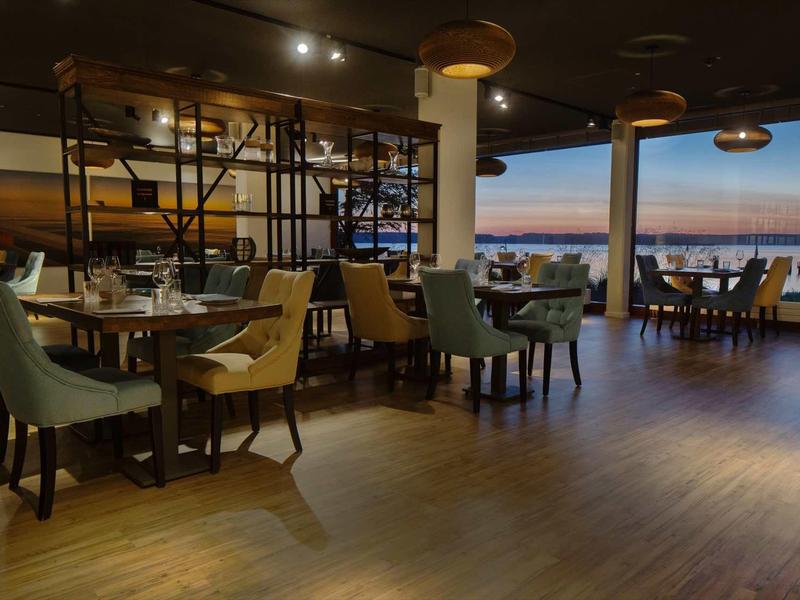 Modernes Restaurant mit Holzboden, bunten Stühlen und großer Fensterfront zum Sonnenuntergang am Meer.