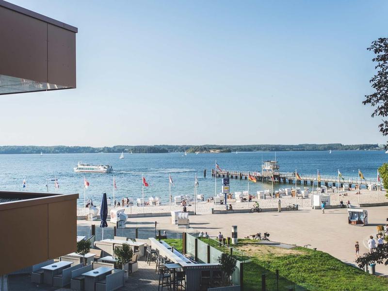 Blick auf den Strand mit vielen Menschen, blauer Himmel, Bäume und moderne Gebäude im Vordergrund.