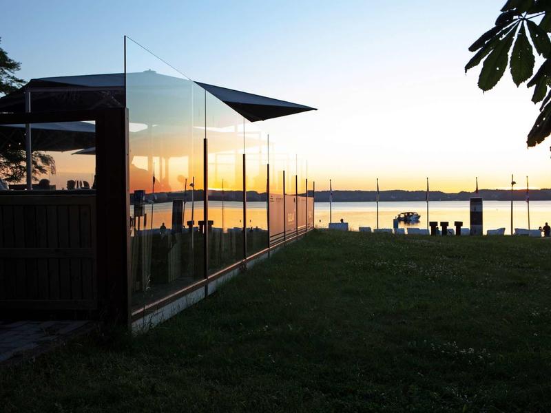 Glashaus am Wasser mit Holzsteg, Wiese im Vordergrund, Sonnenuntergang und klarer Himmel.