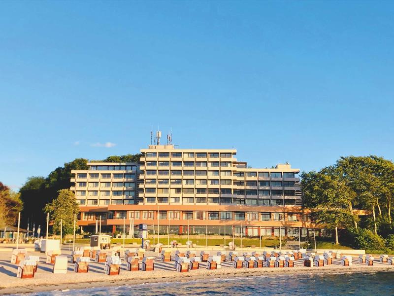 Mehrstöckiges Hotel mit Balkonreihen, davor eine Strandpromenade mit bunten Strandkörben bei blauem Himmel.
