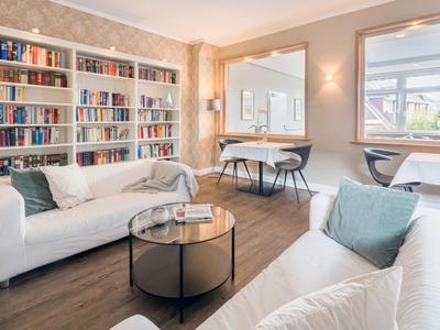 Helles Wohnzimmer mit weißen Sofas, runden Glastisch, großem Bücherregal und Holzboden.