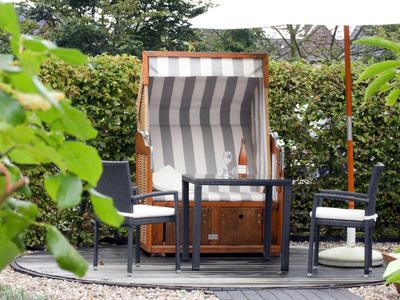 Gemütliche Terrasse mit Strandkorb, zwei Stühlen und einem kleinen Tisch, umgeben von Grün.