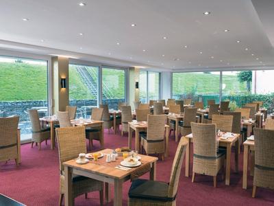 Ristorante luminoso con molti tavoli e sedie, grandi finestre e moquette rossa.