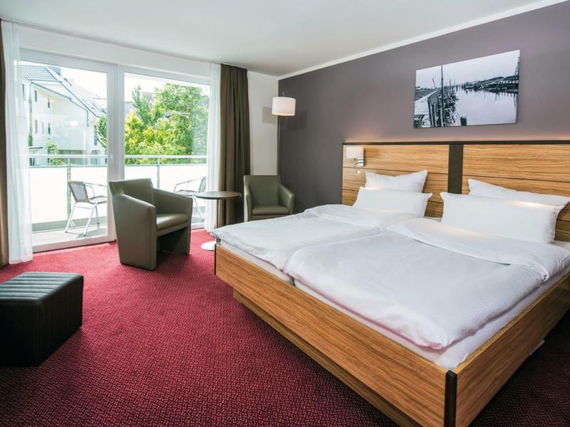 Jasny pokój hotelowy z łóżkiem podwójnym, fotelami i balkonem z widokiem na zieleń.